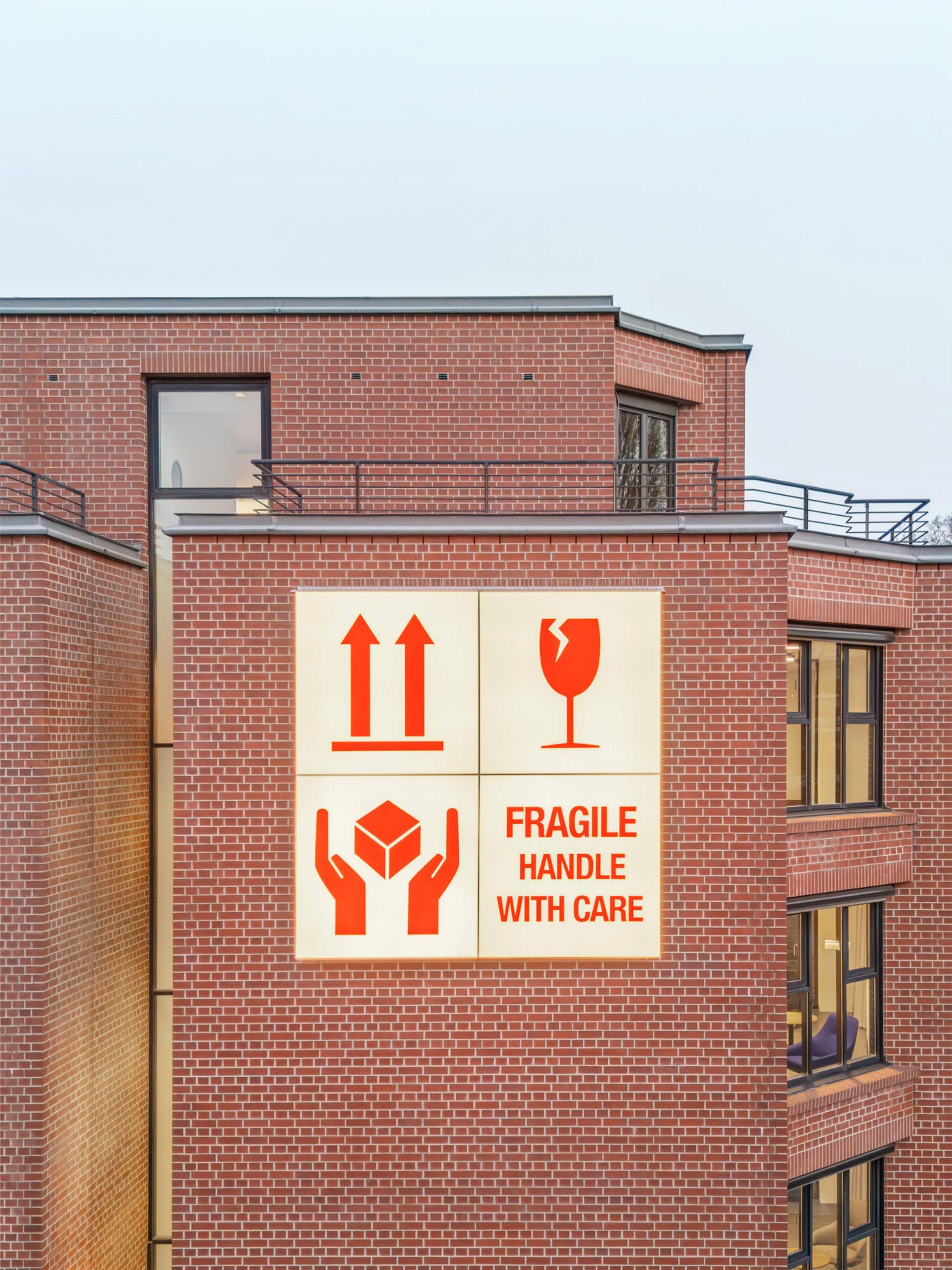 Leuchttafel mit Transportpiktogrammen (Oben, Glasbruch, Vorsicht, Zerbrechlich) an einer Ziegelsteinfassade.
