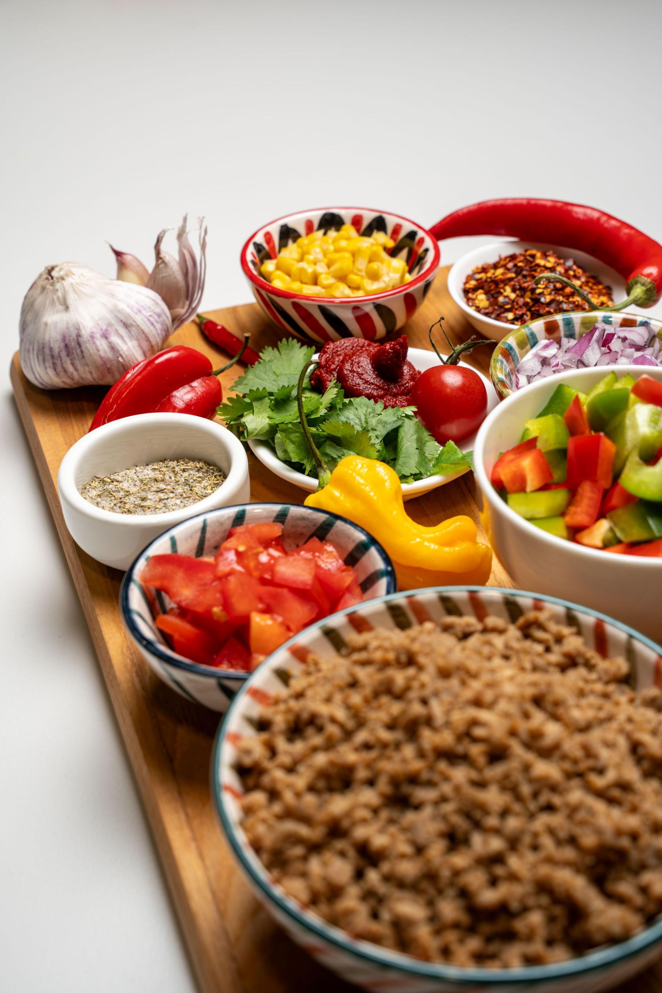 Verschiedene Zutaten wie Hackfleisch, Gemüse, Gewürze und Kräuter für die Zubereitung eines Chili-Gerichts auf einem Holzbrett.