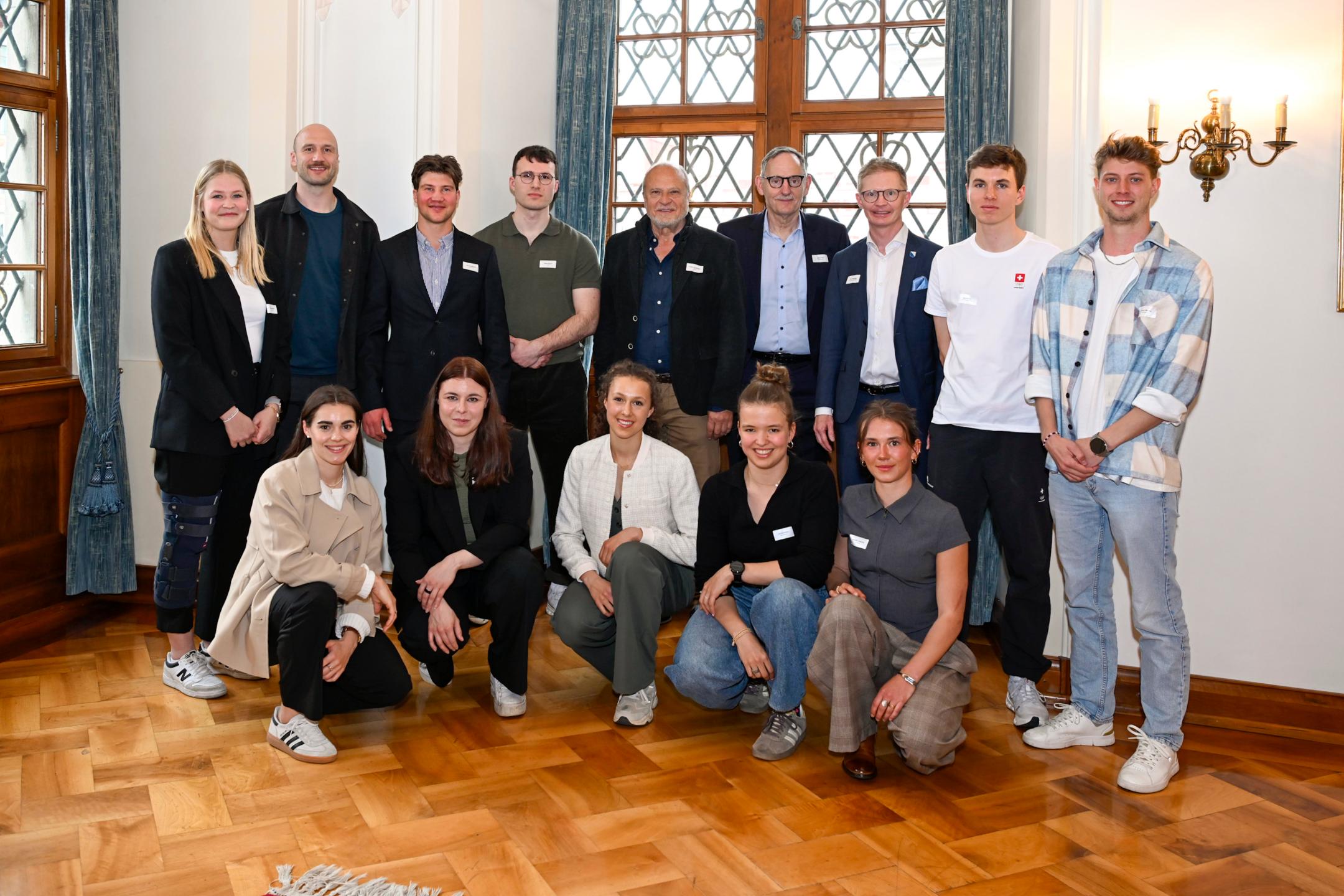 Ein Gruppe sportlicher Menschen in eleganter Kleidung posiert in einem Festsaal zusammen mit drei Männern.