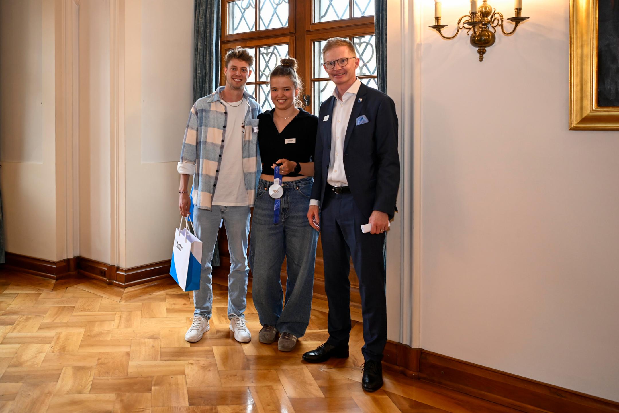 Ein Mann überreicht ein Geschenk an sportliche Frau und einen sportlichen Mann mit Sonnnebrille.