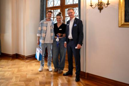 Ein Mann überreicht ein Geschenk an sportliche Frau und einen sportlichen Mann mit Sonnnebrille.