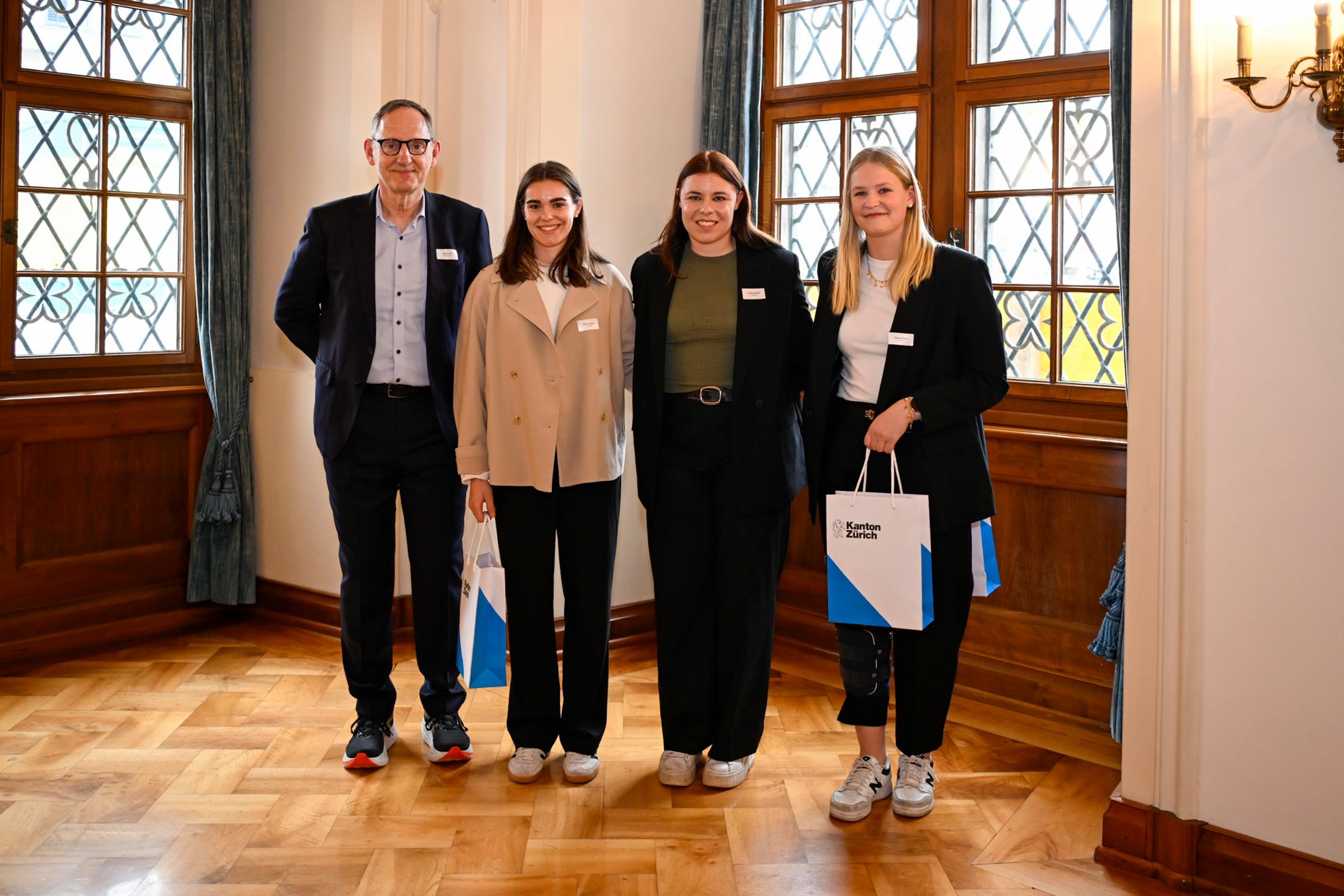 Ein Mann mit Brille und Schnauz überreicht ein Geschenk an drei sportliche Frauen in eleganter Kleidung.