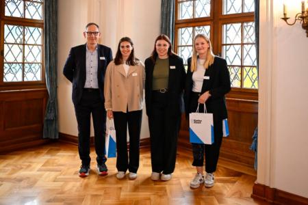 Ein Mann mit Brille und Schnauz überreicht ein Geschenk an drei sportliche Frauen in eleganter Kleidung.
