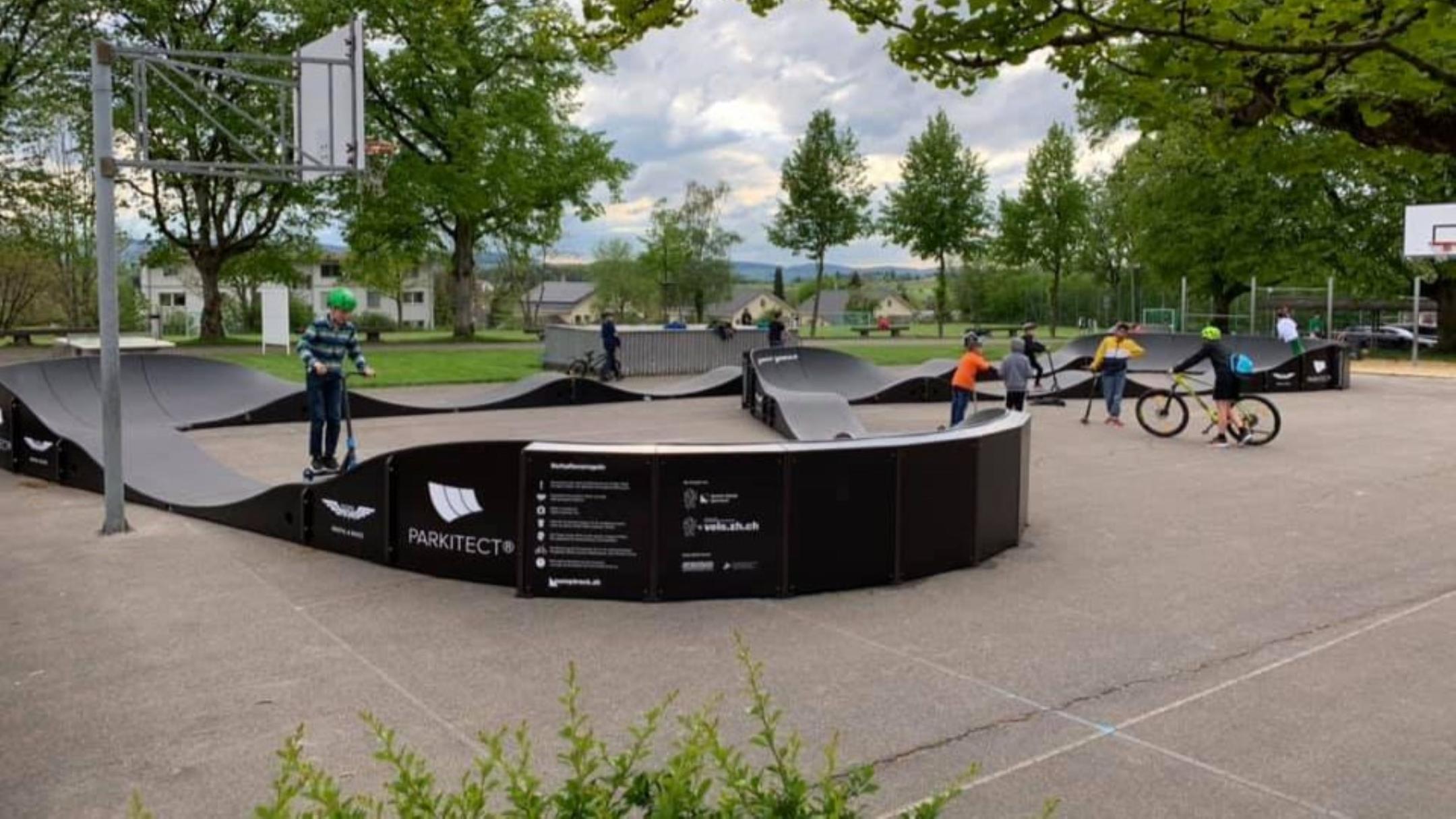 Mehrere Kinder spielen auf einem Pumptrack von «PARKITECT» im Freien. Der Pumptrack hat schwarze Seitenwände und graue Rampen.