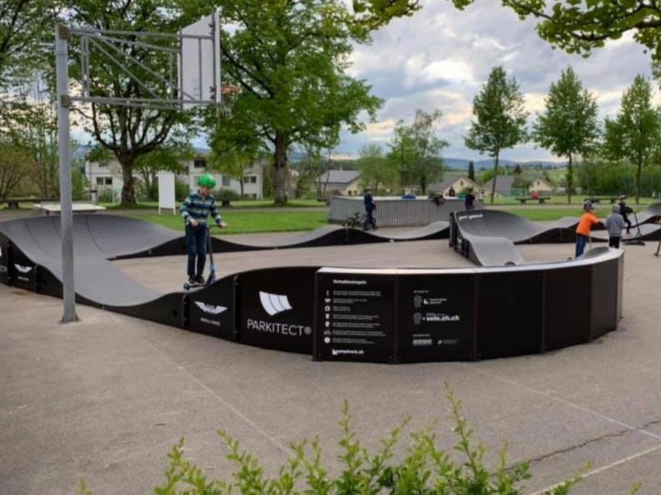 Mehrere Kinder spielen auf einem Pumptrack von «PARKITECT» im Freien. Der Pumptrack hat schwarze Seitenwände und graue Rampen.