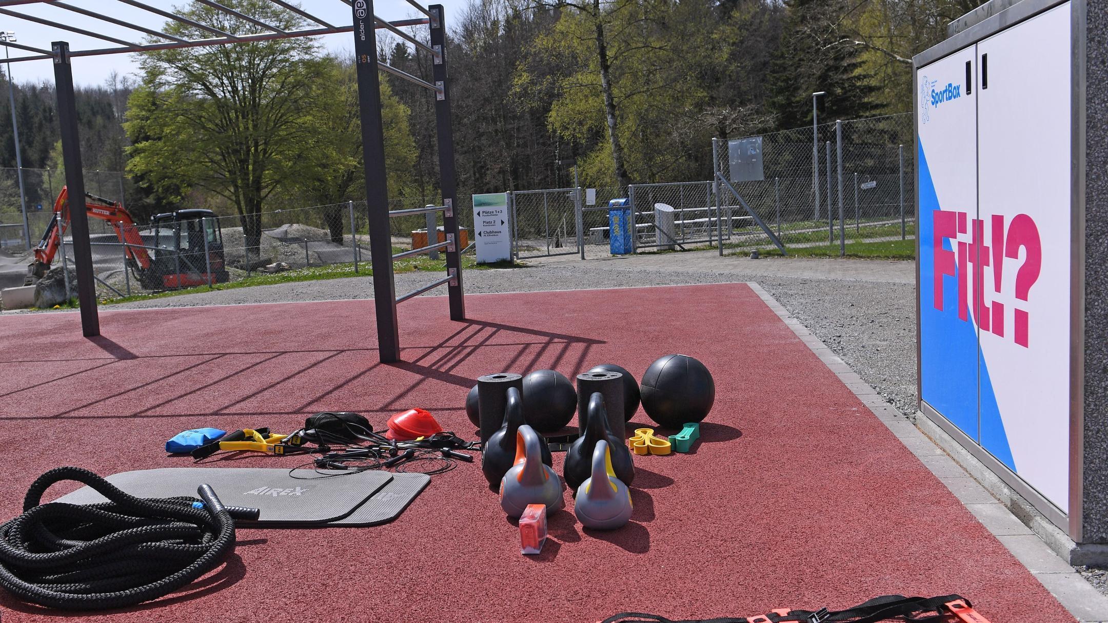 Outdoor-Fitnessbereich mit Trainingsgeräten und einer blauen SportBox mit der Aufschrift «Fit!?».