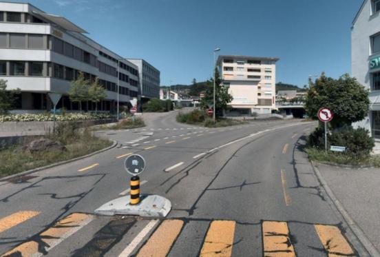 Eine Strasse in einer städtischen Umgebung führt auf eine Kreuzung zu. Im Vordergrund sind gelbe Markierungen eines Fussgängerstreifens und eine kleine Verkehrsinsel zu sehen. Links steht ein mehrstöckiges Bürogebäude mit grauer Fassade, während im Hintergrund und auf der rechten Seite weitere Wohn- und Geschäftshäuser sichtbar sind. Diverse Verkehrsschilder, darunter ein Wendeverbotsschild rechts, säumen die Strasse.