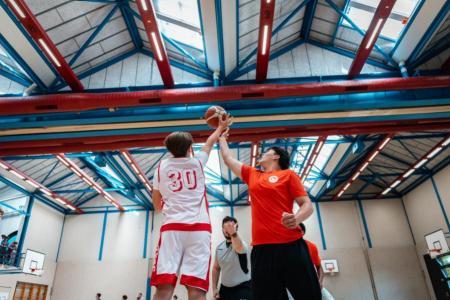 Zwei Knaben spielen Basketball in einer Sporthalle, der Schiedsrichter steht in der Mitte.