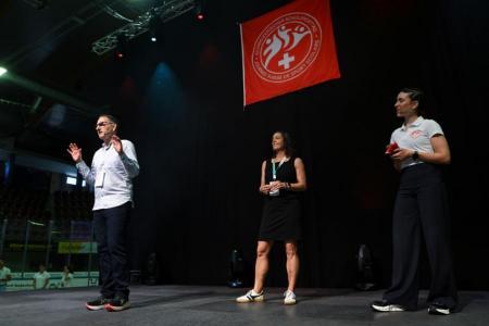 Ein Mann mit weissem Hemd und Brille und eine Frau in schwarzem Kleid und eine Moderatorin in weissem Shirt stehen auf einer Bühne, im Hintergrund eine rote Fahne.
