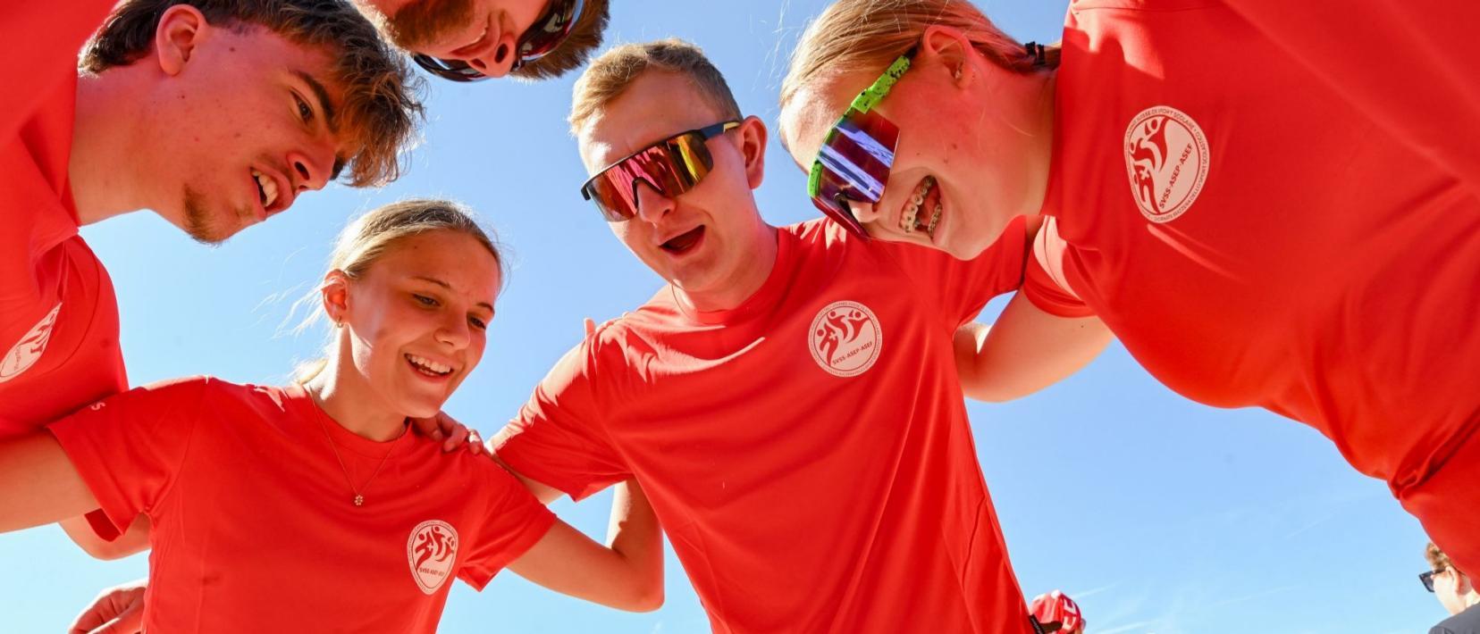 Sechs lachende Jugendliche in rotem T-Shirt bilden einen Kreis und legen die Arme um sich.