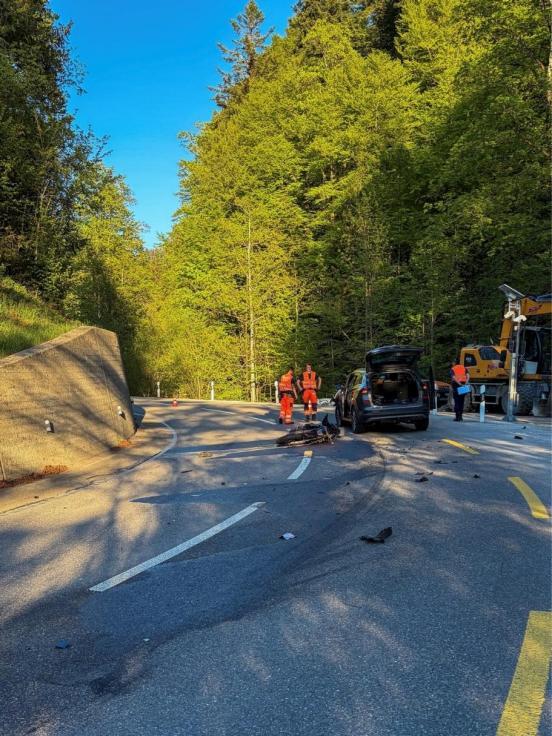 Eine Strasse, die eine links Kurve macht, umgeben von Wald. Ein Töff liegt am Boden neben einem Auto, das den Kofferraumdeckel geöffnet hat. Daneben stehen Polizisten 