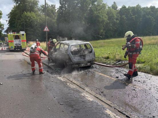 Ein ausgebrannter PW auf einem Trottoir und Feuerwehrleute mit Atemschutz