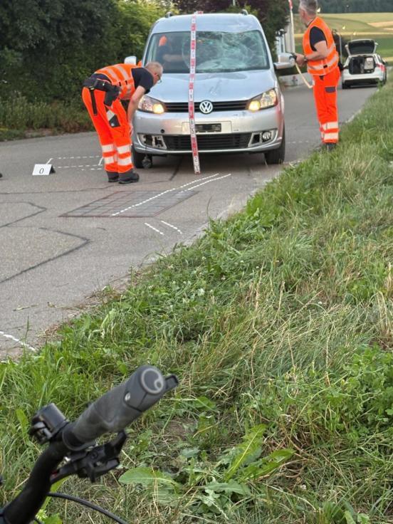 Im Vordergrund die Lenkstange des FAhrrades, im Hintergrund das beteiligte Auto. Zwei Polizisten sichern Spuren.