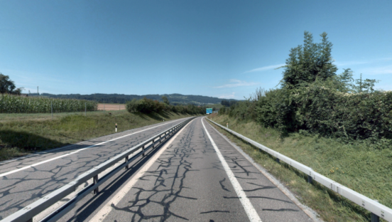 Blick aus der Fahrerperspektive auf eine zweispurige Landstrasse an einem sonnigen Tag unter blauem Himmel. Die Fahrbahn weist deutliche, dunkel versiegelte Risse im Asphalt auf. Links trennt eine doppelte Leitplanke die Gegenfahrbahn ab, rechts verläuft eine einfache Leitplanke entlang eines Grünstreifens mit dichten Büschen. Im Hintergrund führt die Strasse in einer leichten Rechtskurve auf hügeliges, bewaldetes Gelände zu.