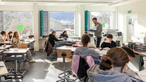 Heller Klassenraum mit mehreren Schülerinnen und Schülern an Gruppentischen. Eine Lehrkraft steht in der Mitte und liest aus einem Buch vor.