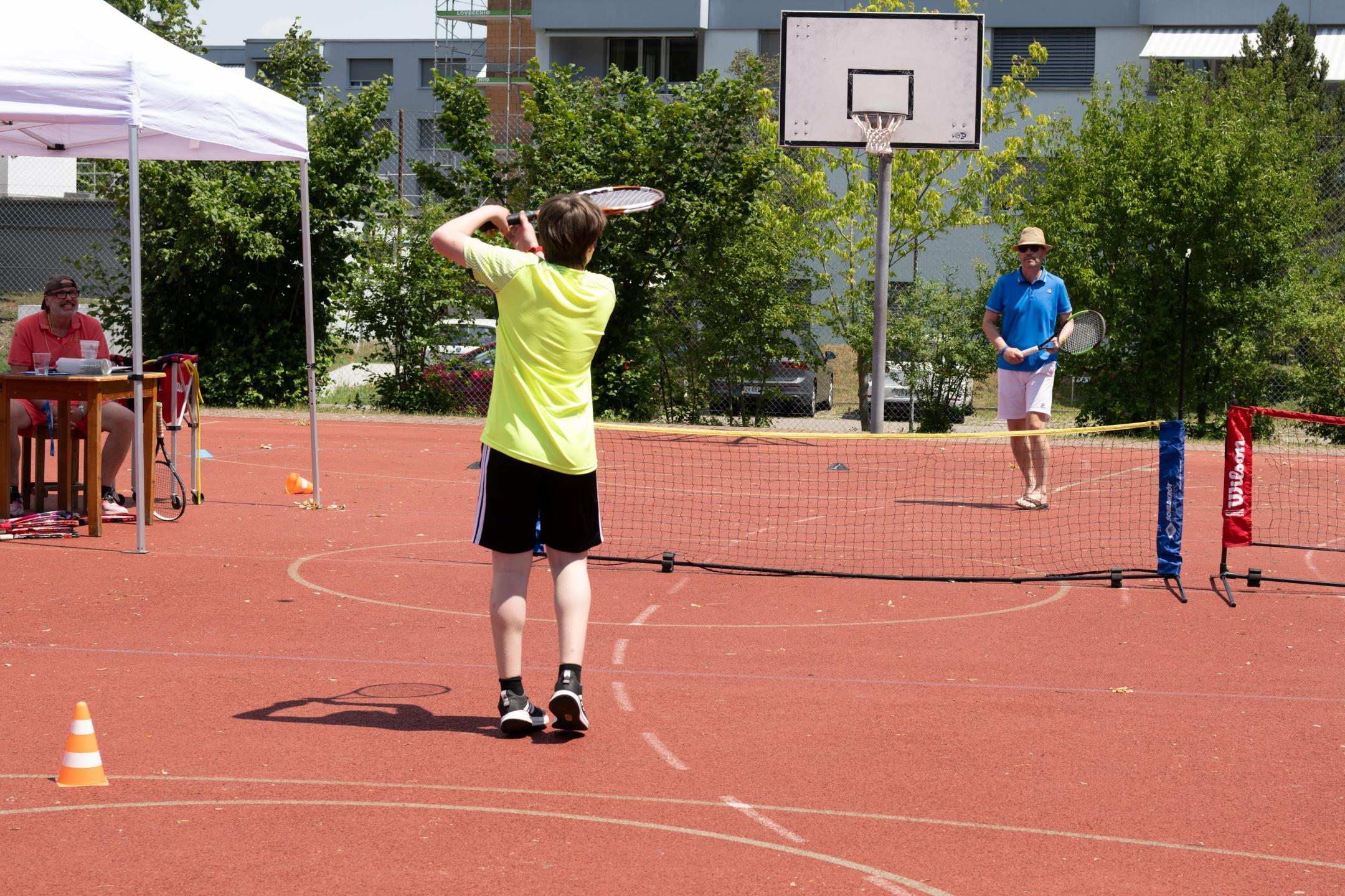 Ein Knabe und ein Mann machen Tennis-Spielformen auf einem roten Platz.