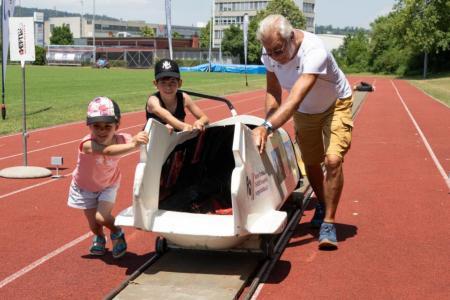 Ein älterer Herr und zwei Kinder schieben einen Bob auf einer Schienen.