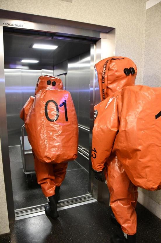 Zwei FOR-Mitarbeitende in organgen Vollschutzanzügen schieben einen Wagen in den Lift