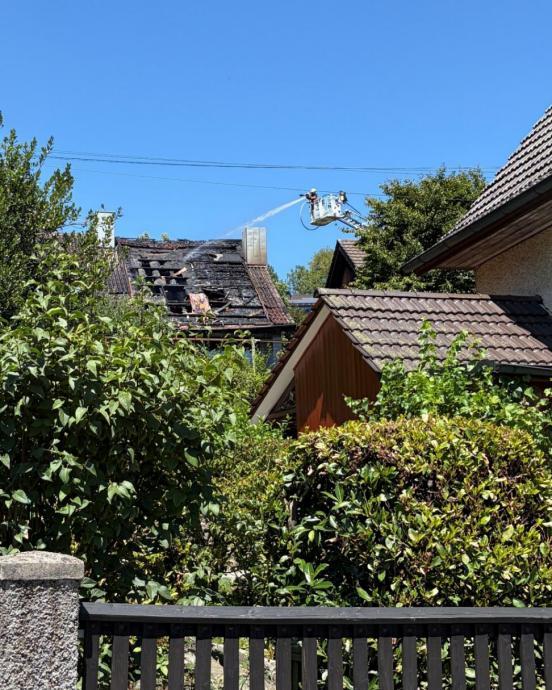 Ein verbranntes Dach eines Einfamilienhauses und zwei Feuerwehrleute die von einer Autodrehleiter aus das Dack abspritzen