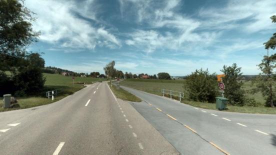 Eine Aufnahme aus der Fahrerperspektive auf eine asphaltierte Strasse. Die Strasse führt durch eine ländliche Landschaft mit grünen Feldern und Wiesen. Rechts zweigt eine kleinere Strasse oder ein Weg ab, der mit einer gestrichelten gelben Linie und einer durchgezogenen weissen Linie markiert ist. Auf der rechten Seite sind Verkehrsschilder zu sehen, darunter ein Wegweiser und ein Verbotsschild für Fahrräder. Im Hintergrund sind Häuser einer Siedlung zu erkennen. Der Himmel ist blau mit einigen Wolken.