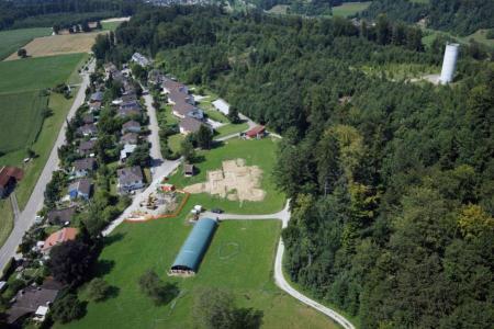 Eine Luftaufnahme zeigt ein Wohngebiet mit Einfamilienhäusern und Gärten neben einem grossen Waldgebiet. Auf einer grünen Wiese sind eine Baustelle und eine halbrunde grüne Halle zu sehen. Ein hoher, weisser Wasserturm ragt am Waldrand empor. Im Hintergrund sind Felder und Hügel erkennbar.