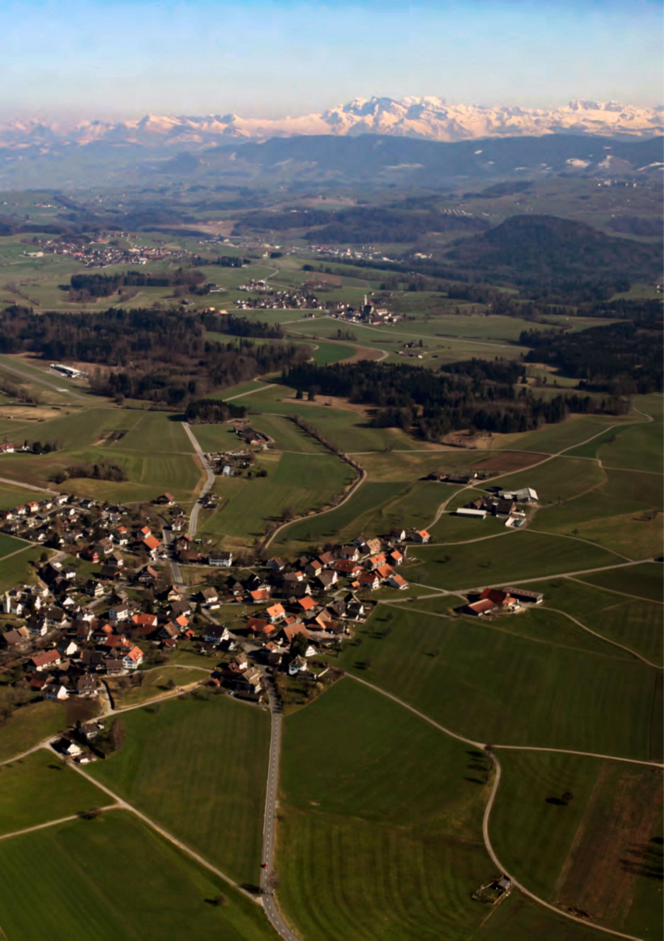 Eine Luftaufnahme zeigt ein Panorama der Schweizer Alpen mit schneebedeckten Gipfeln im Hintergrund und einer grünen Hügellandschaft, Wäldern, Dörfern und links einer Flugplatz-Piste im Vordergrund unter klarem blauem Himmel.