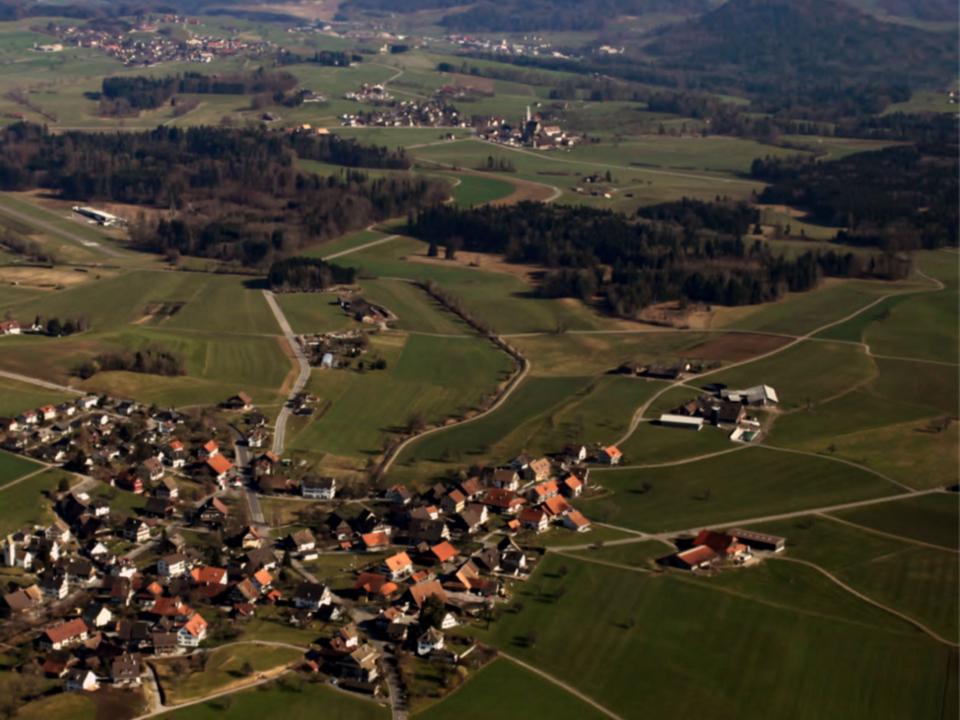 Eine Luftaufnahme zeigt ein Panorama der Schweizer Alpen mit schneebedeckten Gipfeln im Hintergrund und einer grünen Hügellandschaft, Wäldern, Dörfern und links einer Flugplatz-Piste im Vordergrund unter klarem blauem Himmel.