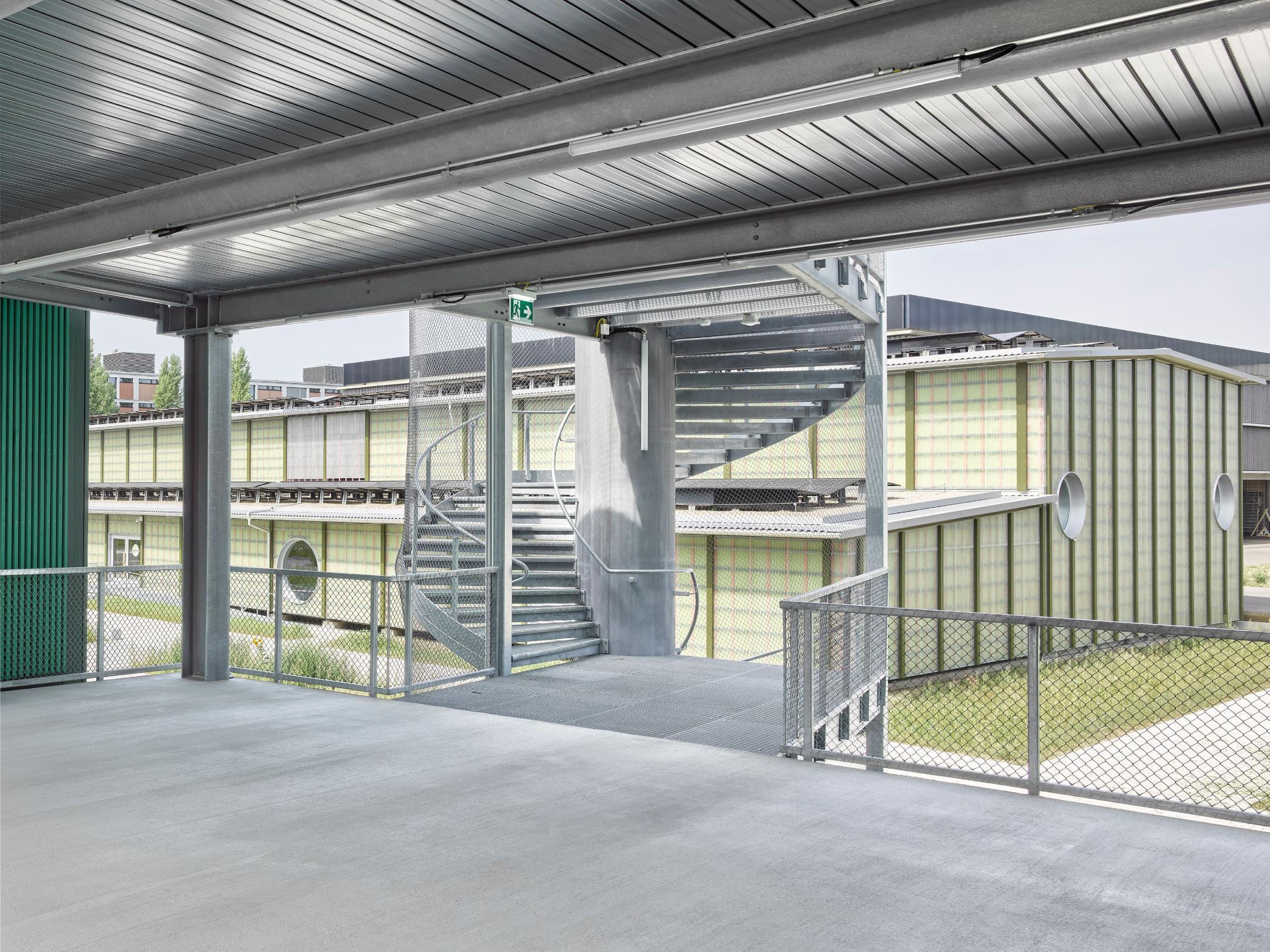 Blick von einer überdachten Betonplattform auf ein Gebäude mit transluzenten Paneelen und einer spiralförmigen Metalltreppe.