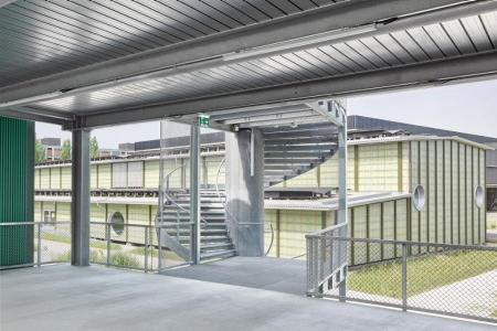 Blick von einer überdachten Betonplattform auf ein Gebäude mit transluzenten Paneelen und einer spiralförmigen Metalltreppe.