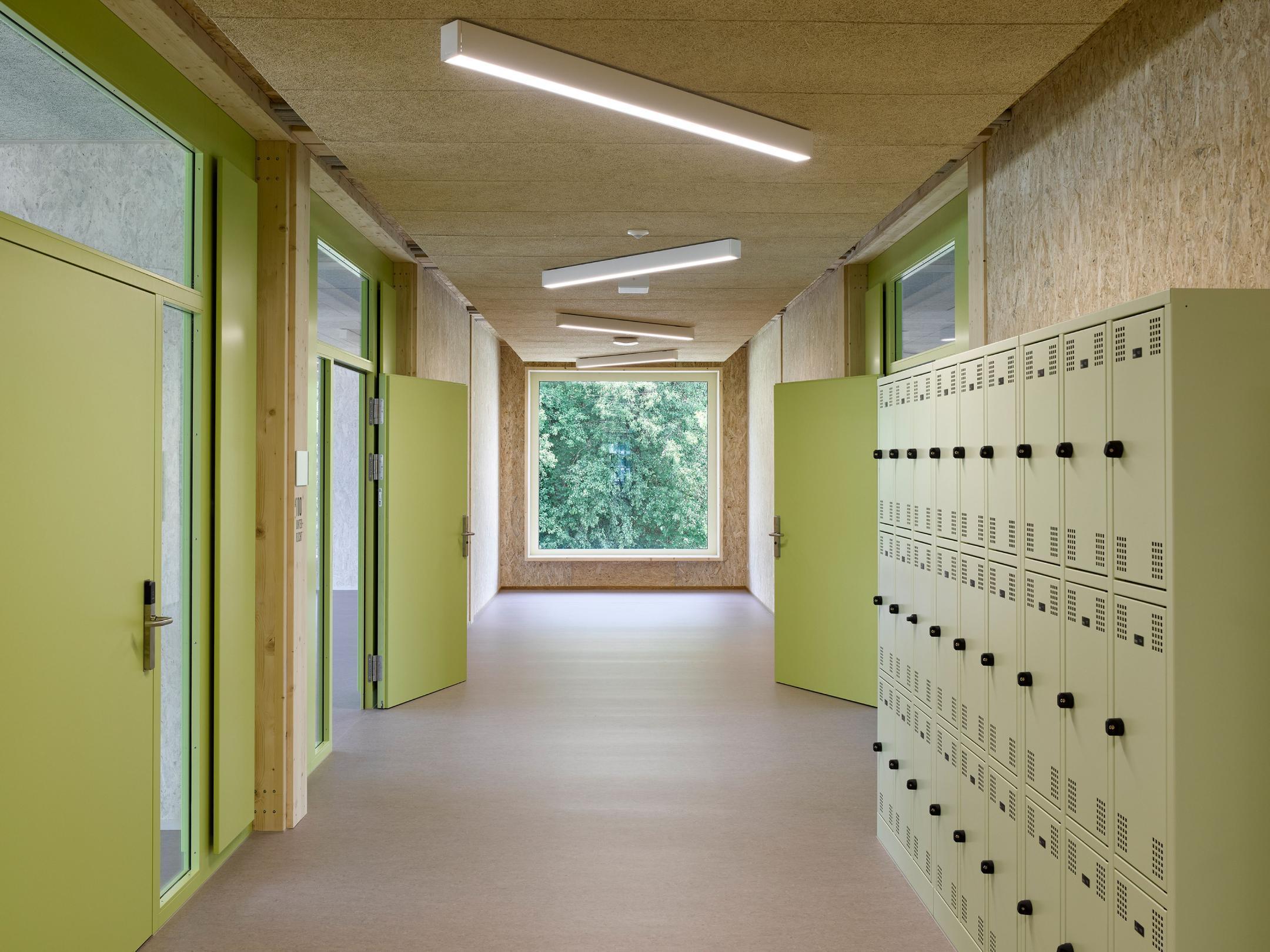 Schulflur mit holzvertäfelten Wänden und Decke, grün gestrichenen Türen und Spinden, und einem Fenster mit Blick ins Grüne am Ende.