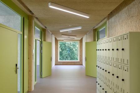 Schulflur mit holzvertäfelten Wänden und Decke, grün gestrichenen Türen und Spinden, und einem Fenster mit Blick ins Grüne am Ende.