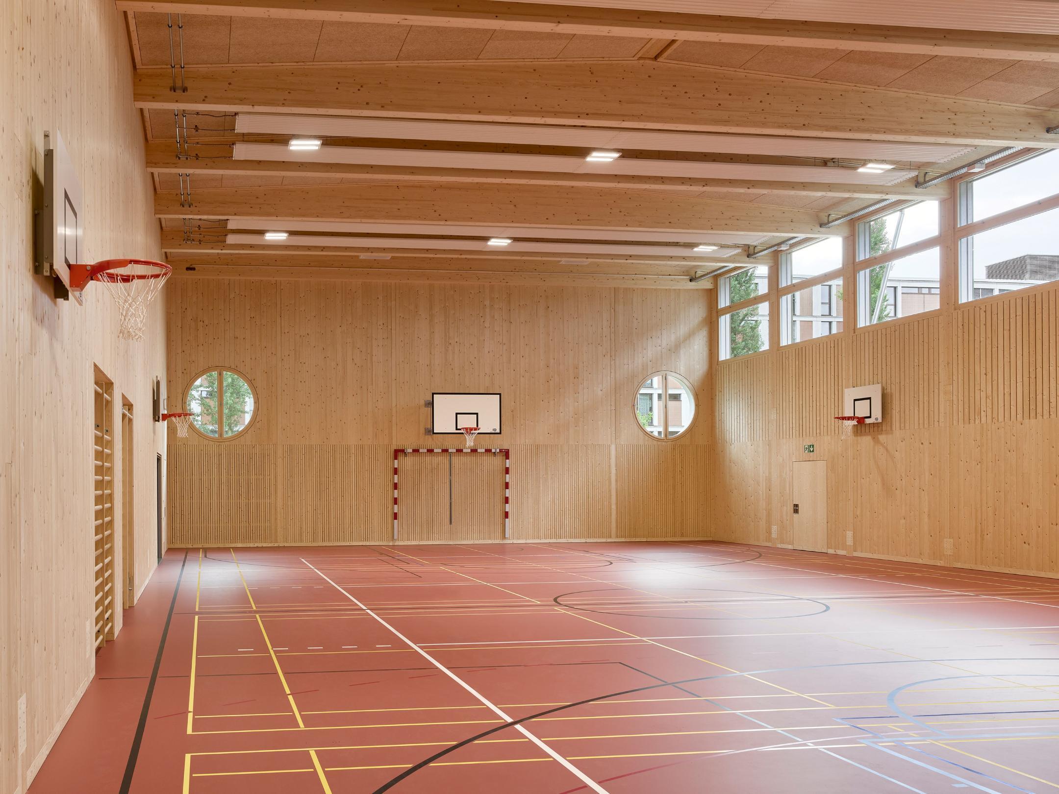 Neue Sporthalle mit hellen Holzwänden, runden Fenstern und rotem Boden mit farbigen Spielfeldmarkierungen. Basketballkörbe und Handballtore sind sichtbar.