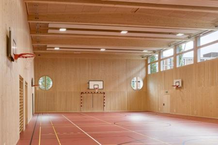 Neue Sporthalle mit hellen Holzwänden, runden Fenstern und rotem Boden mit farbigen Spielfeldmarkierungen. Basketballkörbe und Handballtore sind sichtbar.