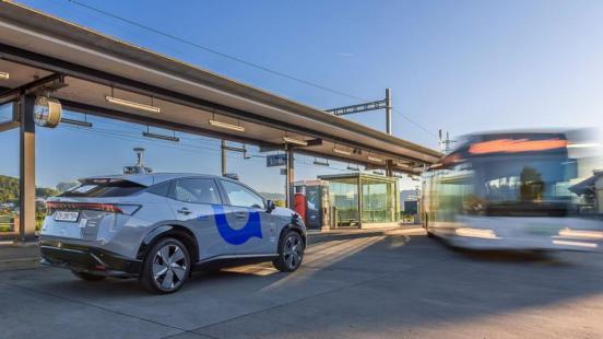 Selbstfahrendes Auto steht am Bahnhof in Otelfingen. Im Hintergrund fährt ein Linienbus von der Haltestelle ab.