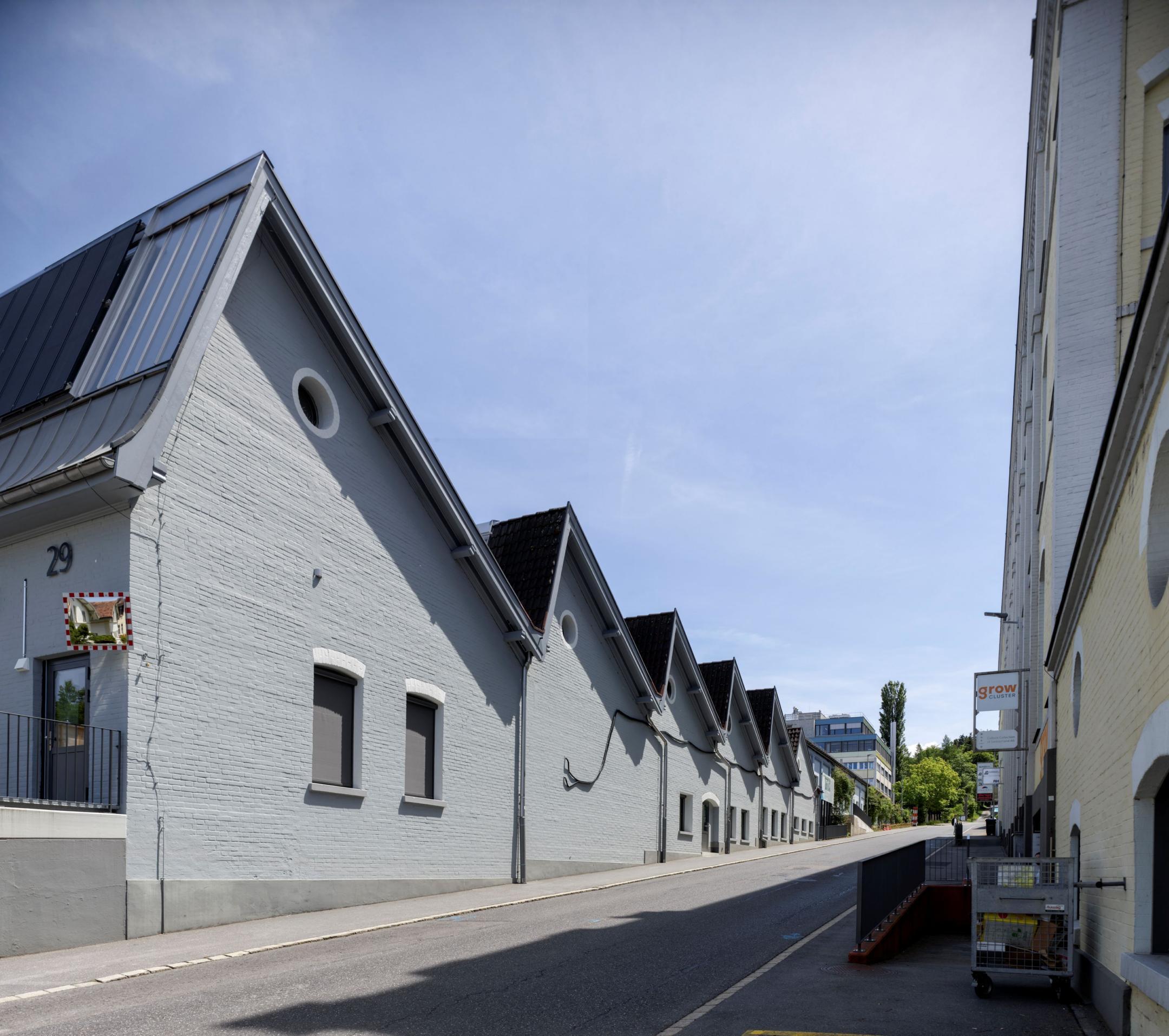 Reihe von grauen Lagerhallen mit gezackten Dächern und runden Fenstern, an einer Strasse. Die Nummer «29» ist an der ersten Halle zu sehen.