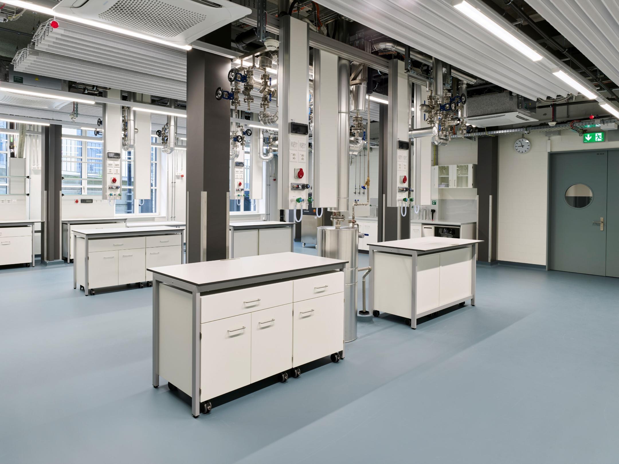 Modernes Labor mit mehreren mobilen Tischen, weissen Schränken und komplexer Rohrinstallation an Decke und Säulen.