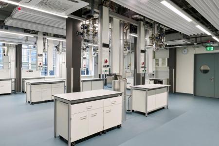 Modernes Labor mit mehreren mobilen Tischen, weissen Schränken und komplexer Rohrinstallation an Decke und Säulen.