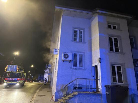 Rechts ein Gebäude, aus dem Rauch aus den Fenstern kommt. Links davon ein Feuerwehrauto.