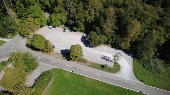 Luftaufnahme eines unbefestigten Kiesparkplatzes am Waldrand. Eine geteerte Strasse verläuft im Vordergrund parallel zum Parkplatz, der von dichten, grünen Bäumen und Wiesen umgeben ist. Auf dem Areal stehen vereinzelt parkierte Autos.