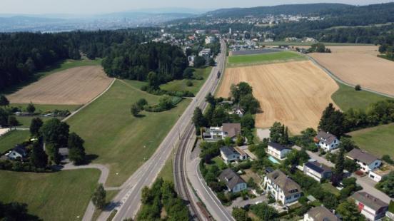 Luftaufnahme des Zollikerbergs mit der zentral verlaufenden Forchstrasse, Bahngleisen, angrenzenden Feldern und Wohnhäusern.