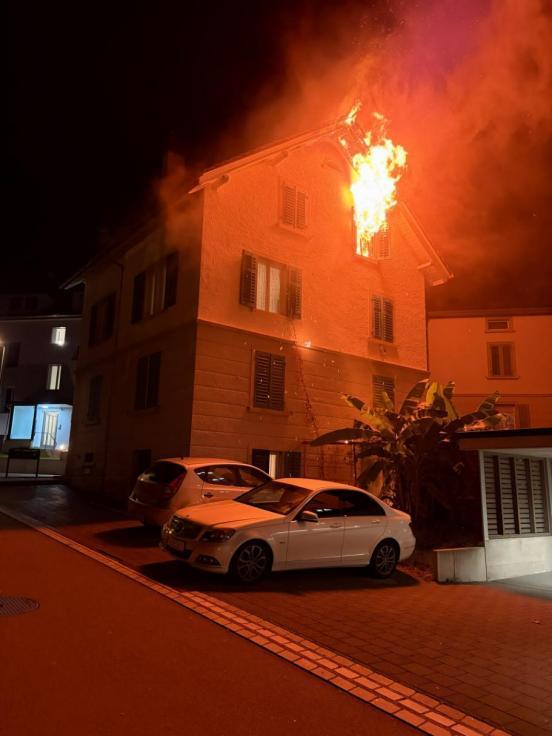 Aus einem Fenster eines Mehrfamilienhauses steigen Flammen empor. Es ist Nacht.