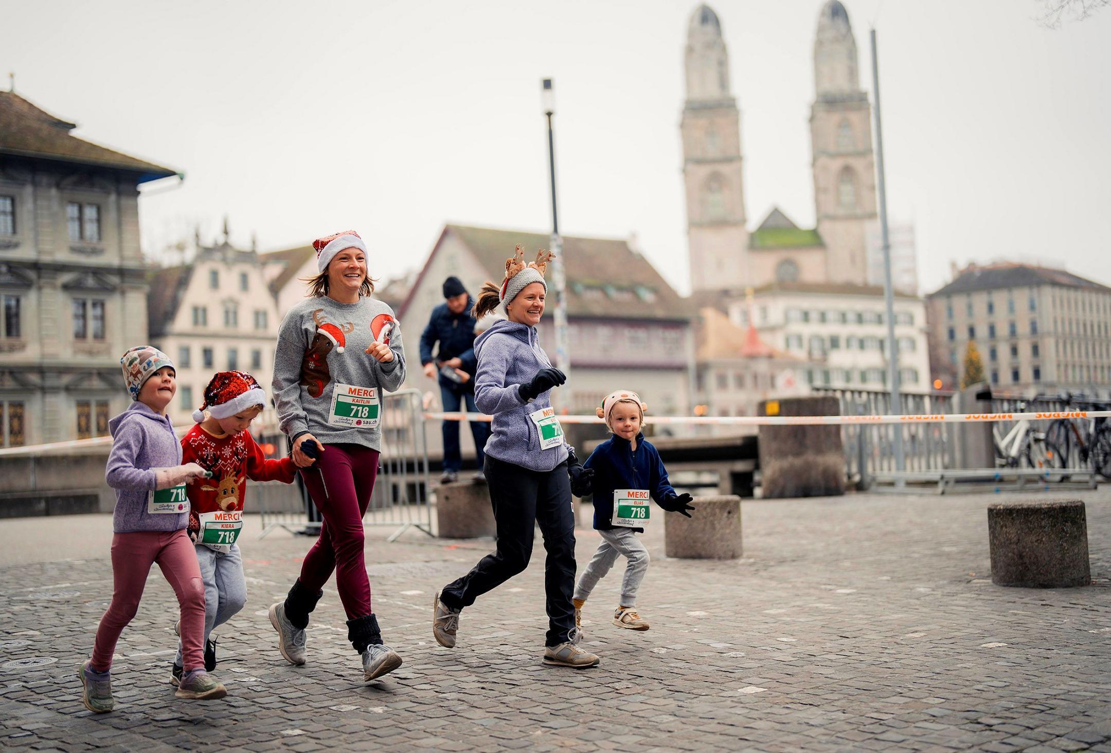 Zwei Läuferinnen mit drei Kindern an der Hand, mit Mützen und Startnummern, rennen am Zürcher Silvesterlauf in der  Innenstadt. 