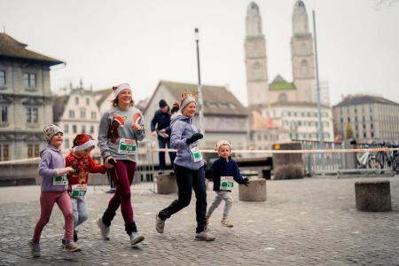 Zwei Läuferinnen mit drei Kindern an der Hand, mit Mützen und Startnummern, rennen am Zürcher Silvesterlauf in der  Innenstadt. 
