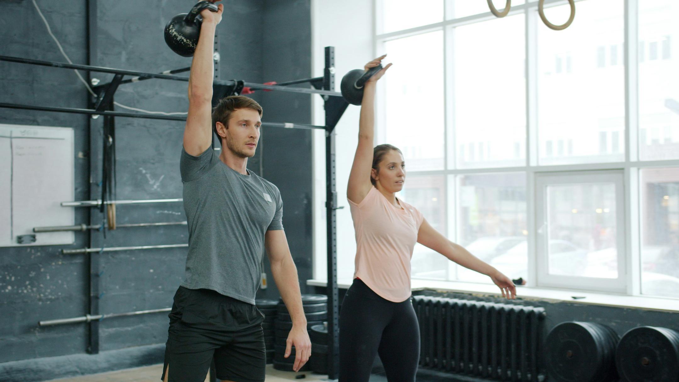 Ein Mann und eine Frau in Sportbekleidung heben einarmig Gewichte in einem Fitnesscenter.