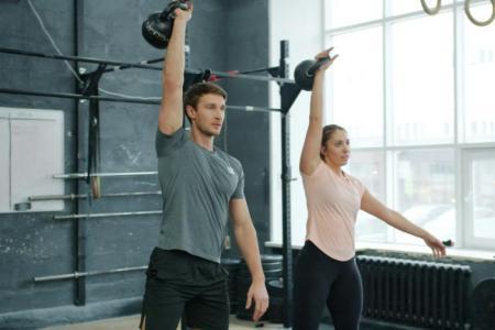 Ein Mann und eine Frau in Sportbekleidung heben einarmig Gewichte in einem Fitnesscenter.