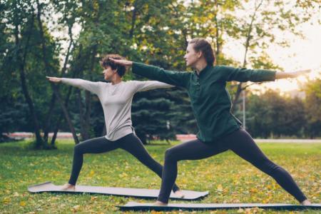 Zwei Frauen in Sportbekleidung stehen auf einem Park in einer Yogapose auf einer Matte.