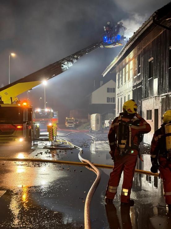 Rechts vorne zwei Feuerwehrleute, links ein Drehleiterfahrzeug, im Hintergurund das rauchende Gebäude