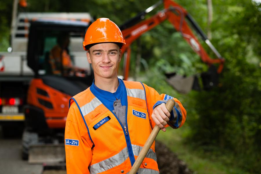 Ein Fachmann Betriebsunterhalt EFZ, Werkdienst, in oranger Arbeitskleidung bei der Arbeit. Hinter ihm ein roter Bagger.