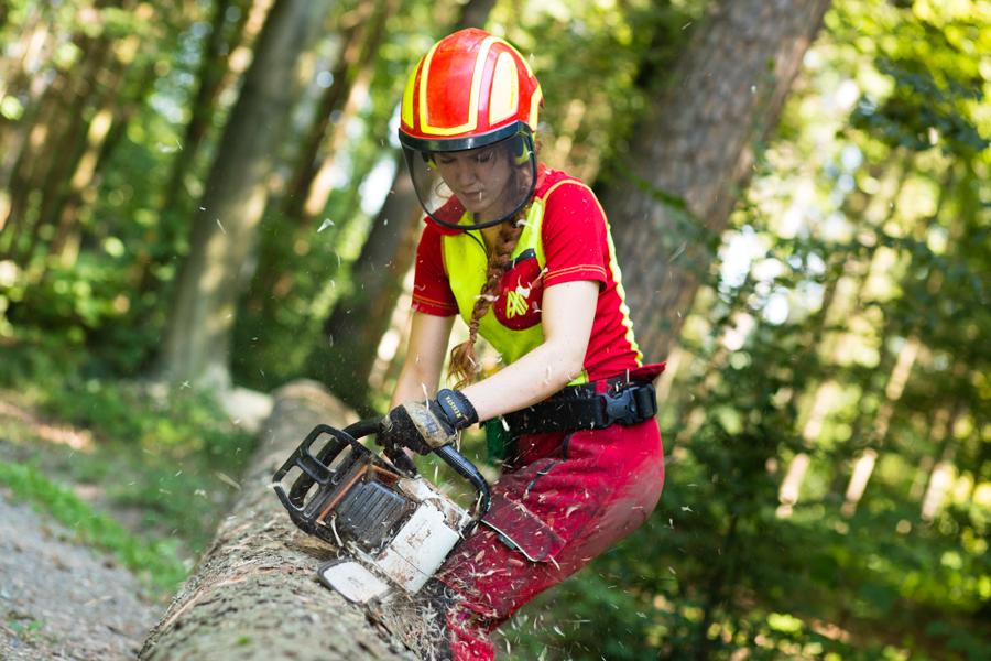 Eine Forstwartin mit Schutzhelm und Arbeitskleidung zersägt im Wald einen Baumstamm mit einer Motorsäge. 