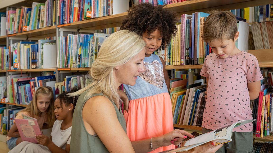 Frau liest zwei Kindern in einer Bibliothek ein Buch vor